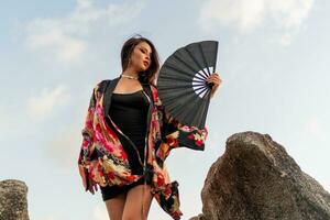 sensual asiático mujer en seda kimono participación ventilador y posando terminado rocas en el playa. foto