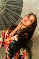 Seductive japanese woman in stylish  kimono  with large fan and professional make up posing over rocks. photo