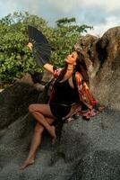 Sensual asian woman in silk kimono  holding fan and posing over rocks on the beach. photo