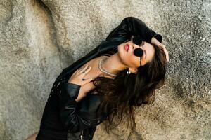Confident brunette woman  in trendy black leather jacket and sensual black dress posing on rocky background. photo