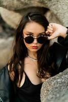 Close op portrait of confident woman  with red lips  in trendy black leather jacket and sensual black dress posing on rocky background. photo