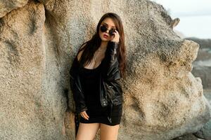 Fashionable  sensual woman in black leather jacket  and stylish sunglasses posing over rocks. photo