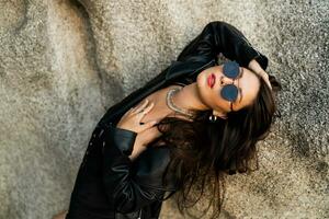 Confident brunette woman  in trendy black leather jacket and sensual black dress posing on rocky background. photo