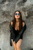 Gorgeous asian woman  with red lips  in trendy black leather jacket and sensual black dress posing on rocky background. photo