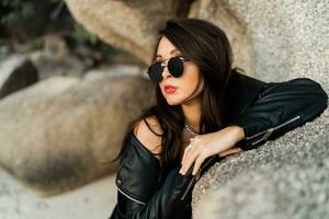 Close op portrait of confident woman  with red lips  in trendy black leather jacket and sensual black dress posing on rocky background. photo