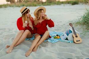 Two pretty women in red summer outfit abd straw hats enjoing picnic on the beach. Summer mood. photo