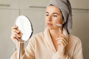 Self Care Concept. Portrait Of Attractive Young Female Looking At Mirror enjoying Her Reflection,  using face massage roller. photo