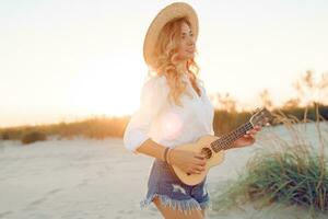 Shapely ginger lady playing ukulele guitar  in sun light on the beach . Wearing straw hat and trendy jeans shorts. Warm sunset colors. photo