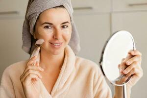 Self Care Concept. Portrait Of Attractive Young Female Looking At Mirror enjoying Her Reflection,  using face massage roller. photo