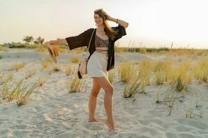 Summer photo of happy young woman in stylish boho  outfit  holding straw bag and hat  posing on  the beach.  Sunset. Full lenght.