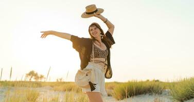 Summer photo of happy young woman in stylish boho  outfit  holding straw bag and hat  posing on the l beach. Full lenght.