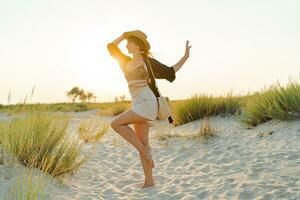 contento europeo mujer en elegante boho verano vestir caminando en el playa. calentar puesta de sol colores.paja sombrero. foto