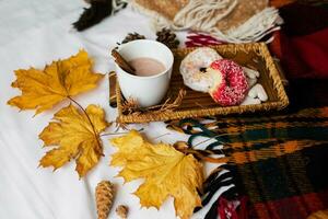 cozy autumn image of tasty breakfast in bed on wooden tray with cup of cacao, cinnamon, cookies and glazed donuts.Warm toned colors, top view, yellow leaves and cones. photo