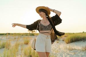 Summer photo of happy young woman in stylish boho  outfit  holding straw bag and hat  posing on the l beach. Full lenght.