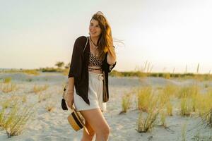 Summer photo of happy young woman in stylish boho  outfit  holding straw bag and hat  posing on  the beach.  Sunset.