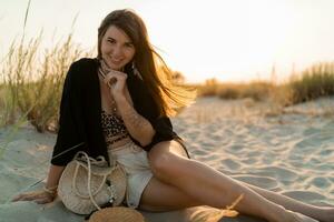 Cheerful brunette woman in stylish summer  boho outfit posing on the beach.  Sunset colors. photo