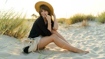 Cheerful brunette woman in stylish summer  boho outfit posing on the beach.  Sunset colors. Straw hat. photo