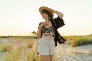 Summer photo of happy young woman in stylish boho  outfit  holding straw bag and hat  posing on the l beach. Full lenght.