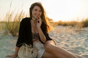 alegre morena mujer en elegante verano boho atuendo posando en el playa. puesta de sol colores. foto