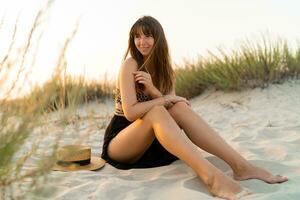 sexy mujer en elegante verano boho atuendo sentado en arena y disfrutando vacaciones en el playa. puesta de sol colores. foto