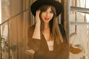 Close up autumn I portrait of elegant woman in black hat and blouse posing in bohemian interior with stairs. photo