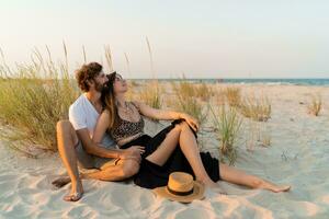 elegante Pareja en amor posando en el playa. morena mujer en Paja sombrero con su novio Relajado en calentar verano noche. foto