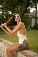 Outdoor summer  fashion photo of elegant seductive woman with wavy hairs posing on  tropical beach while summer vacations. Wearing white stylis body swimwear.