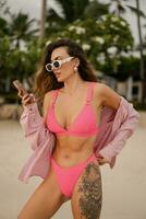 Beautiful stylish woman in  trendy pink swimwear posing on the beach. Summer vacation, tropical island,  palmtrees.  Wearing casual sunglasses. photo