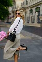 Fashion outdoor photo of blond woman in  white blouse and silk skirt , holding  pink boquet with pions flowers.  Stylish hand bag and sunglasses.  Full lenght.