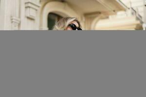 Beautiful smiling blond woman in elegant luxury sunglasses posing on the street. European background. Perfect wavy hairs. photo