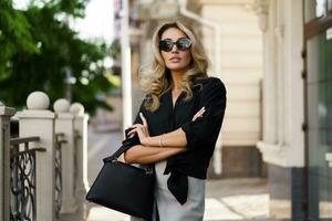 al aire libre retrato de hermosa rubio mujer en Gafas de sol con Perfecto ondulado pelos participación lujo mano bolsa. Moda accesorios , casual atuendo. foto