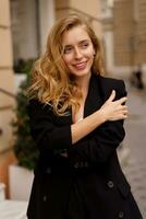 hermosa sonriente mujer en elegante negro traje posando al aire libre en antiguo europeo ciudad. rubio ondulado pelos, perfecto piel. foto