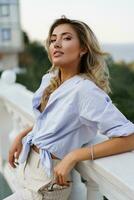 Beautiful  charming blond woman with perfect skin and wavy hairs  posing outdoor during vacation in  Europe.  Wearing blue blouse and white pants.  Summer fashion. photo
