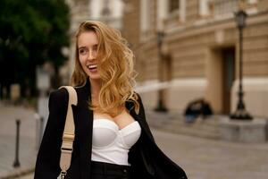 hermosa sonriente mujer en elegante negro traje posando al aire libre en antiguo europeo ciudad. rubio ondulado pelos, perfecto piel. foto