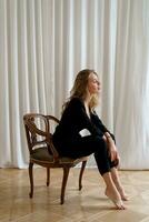 Indoor studio photo of elegant blond woman sitting on the chair. Casual  fashion look. Elegance, seductive mood.
