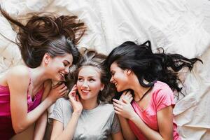 Top view of three best friends tell secrets in  bed . Group of pretty  young  women  in pajamas  lie on back and  talk.  Sunny warm colors. Cozy atmosphere. photo