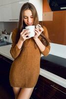 Attractive young  pretty  woman with white cup of tea looking out of the camera in the modern kitchen on sunny fall morning time. photo
