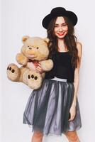 Close up  portrait of pretty cute young woman in stylish hipster outfit  with big eyes , red lips and perfect skin  holding her fluffy teddy bear against white urban wall. photo