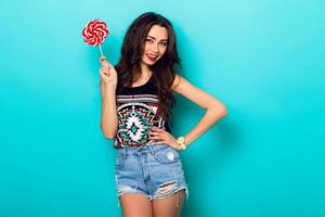 Studio closeup colorful portrait of young sexy funny fashion girl posing on blue wall background in summer style outfit with red lollipop wearing blue jeans and boho t-short. photo