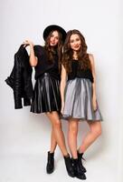 fashion portrait of two elegant stylish women wearing a leather skirt and black hat. Posing against white background.They smiling and looking at camera. photo