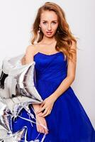 Pretty young women celebrate  birthday  and holding  silver party balloons. Wearing blue evening stylish  dress and have day of against white wall. photo