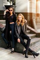 dos elegante bonito muchachas posando en el calle en soleado día. vistiendo de moda urbano atuendo , cuero chaqueta y botas tacones foto