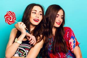 Close up  lifestyle portrait of two  attractive  fancy  friends  in  colorful hipster  fashionable   outfit .  Women  holding pink lolly pop and having fun together against blue wall photo