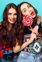 Close up  lifestyle portrait of two  attractive  cute   friends  in  colorful geometric  print    outfit .  Young model  holding a large pink lollipop delicious and laughing against blue wall. photo