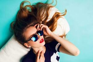 cerca arriba Moda retrato de bonito juguetón dama en elegante Gafas de sol, baloncesto t-corto y blanco zapatillas sentado en blanco almohada en contra brillante agua antecedentes . foto