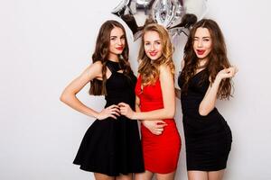 Three best friends celebrate  birthday indoor wearing elegant evening dress and have bright make up. Girls hugging and Showing signs with their hands. Looking at camera and smiling. Inside. photo