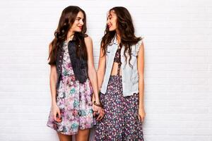 Close up   portrait of two pretty hipster  sisters  wearing boho colorful dress, stylish jeans jacket  and  baubles. Girls smile, have fun against  urban white wall. photo