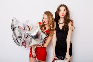 Two beautiful elegant women with red lips in evening black and red dress having fun. One keeping silver stars balloons in her hand . Two woman at the party. Inside. White background. photo