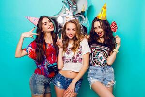 Three beautiful  happy sexy woman  in  stylish summer outfit , paper hats and purity balloons having fun and celebrate  birthday.  Colorful blue background . Pretty girl  hold big lollypop. photo