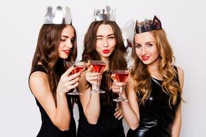 Three beautiful elegant women celebrate hen-party and drinking cocktails. Best friends wearing black stylish evening dress, high heel shoes ,crown on head .Bright make up, red lips. Inside. photo
